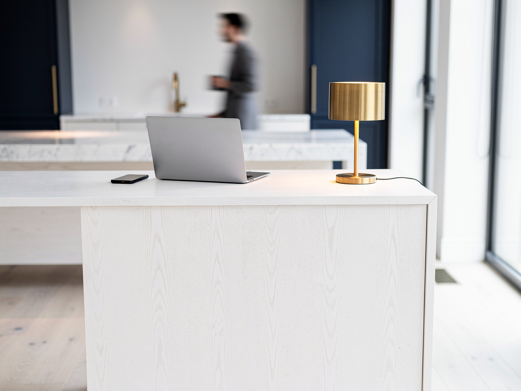 A minimalist front desk in white oak with a laptop, a phone and a small brass lamp, a blurred figure in the background.
